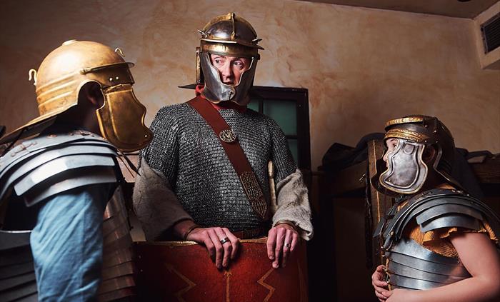 A gladiator stands in the middle of the room. He is flanked by two children trying on Roman armour and helmets. The gladiator explains what it was lik